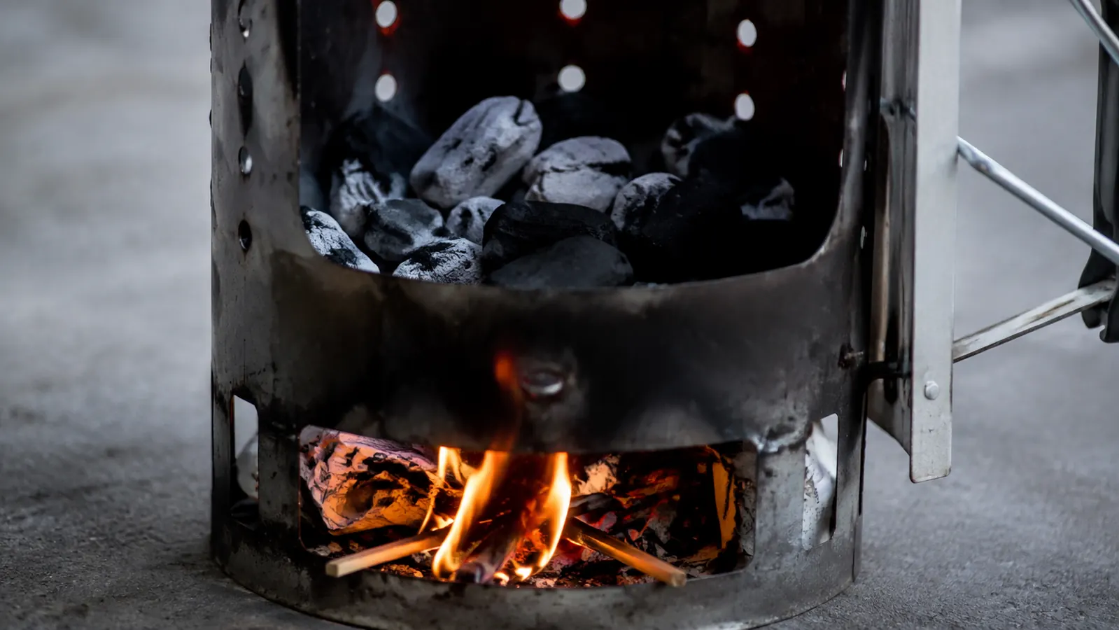 A close view of a chimney starter with wadded newspaper at the base, thin flames licking up, coals beginning to gray