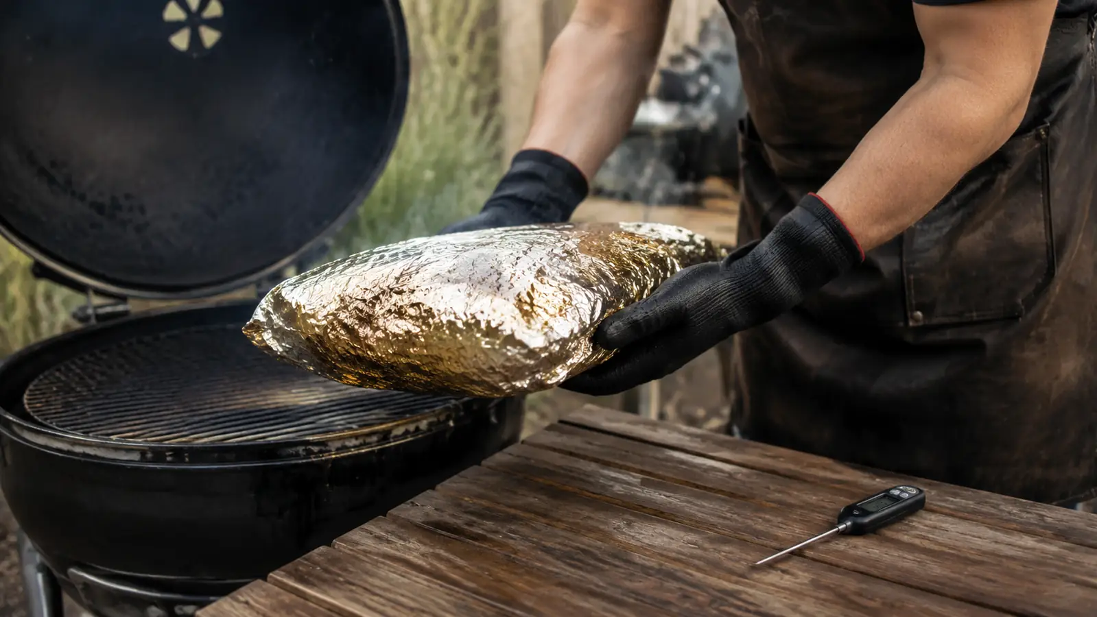 Gloved hands lifting a large foil-wrapped brisket off a smoker rack onto a wooden patio table, evening light