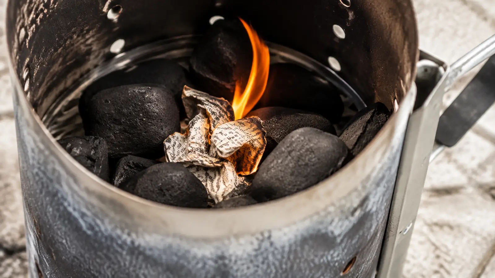 A close look at crumpled newspaper and charcoal in the base of a chimney starter as the fire catches cleanly from underneath.