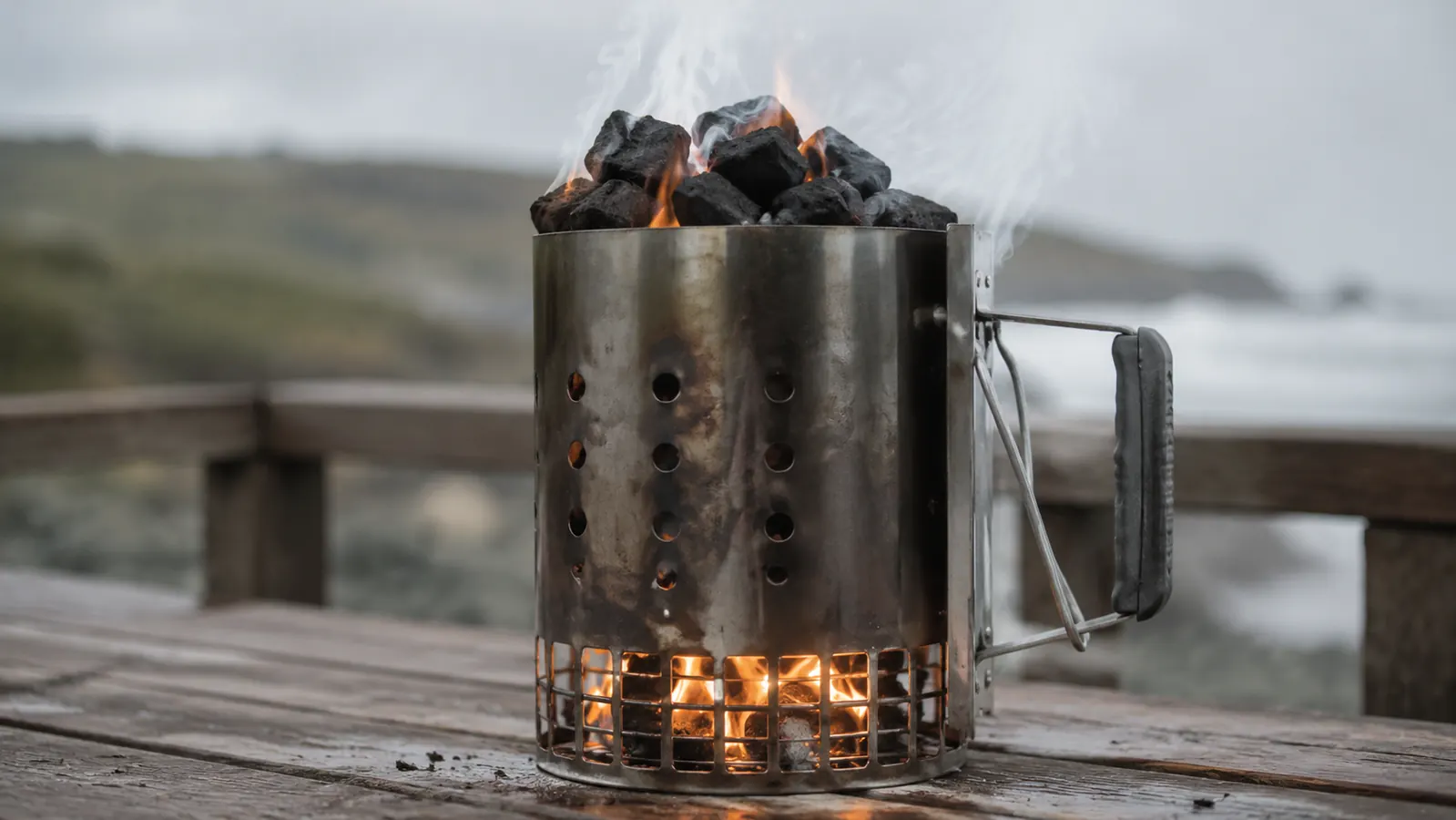 Newspaper flames rise through the bottom vents of a chimney starter on a wooden deck table, morning light catching the column of charcoal.