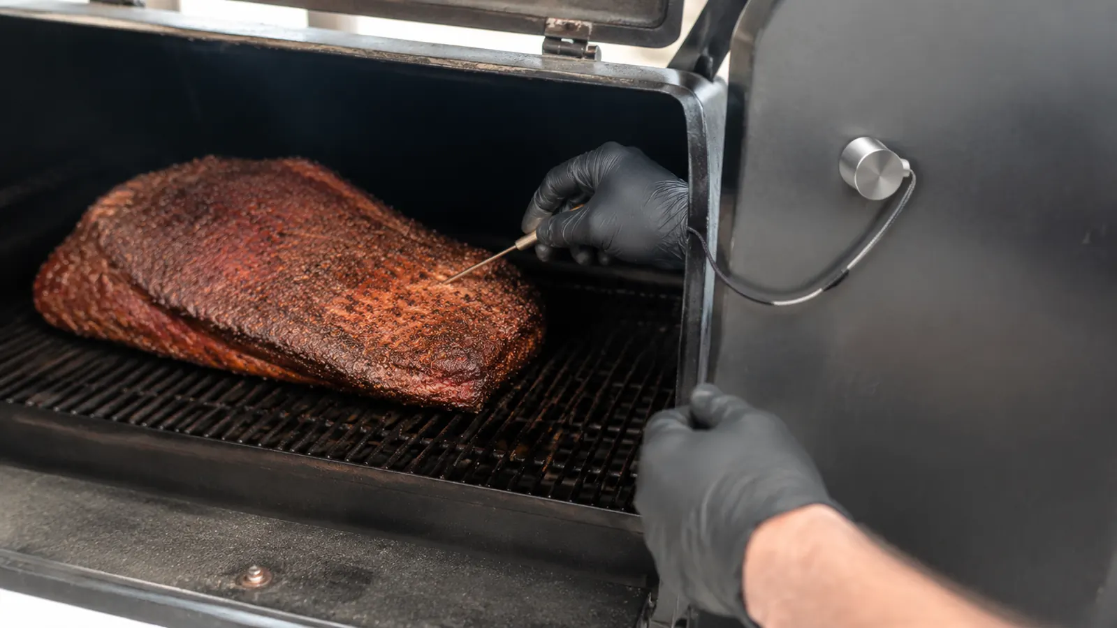 A heat-gloved hand sliding a wireless probe skewer into a brisket resting on the smoker grate